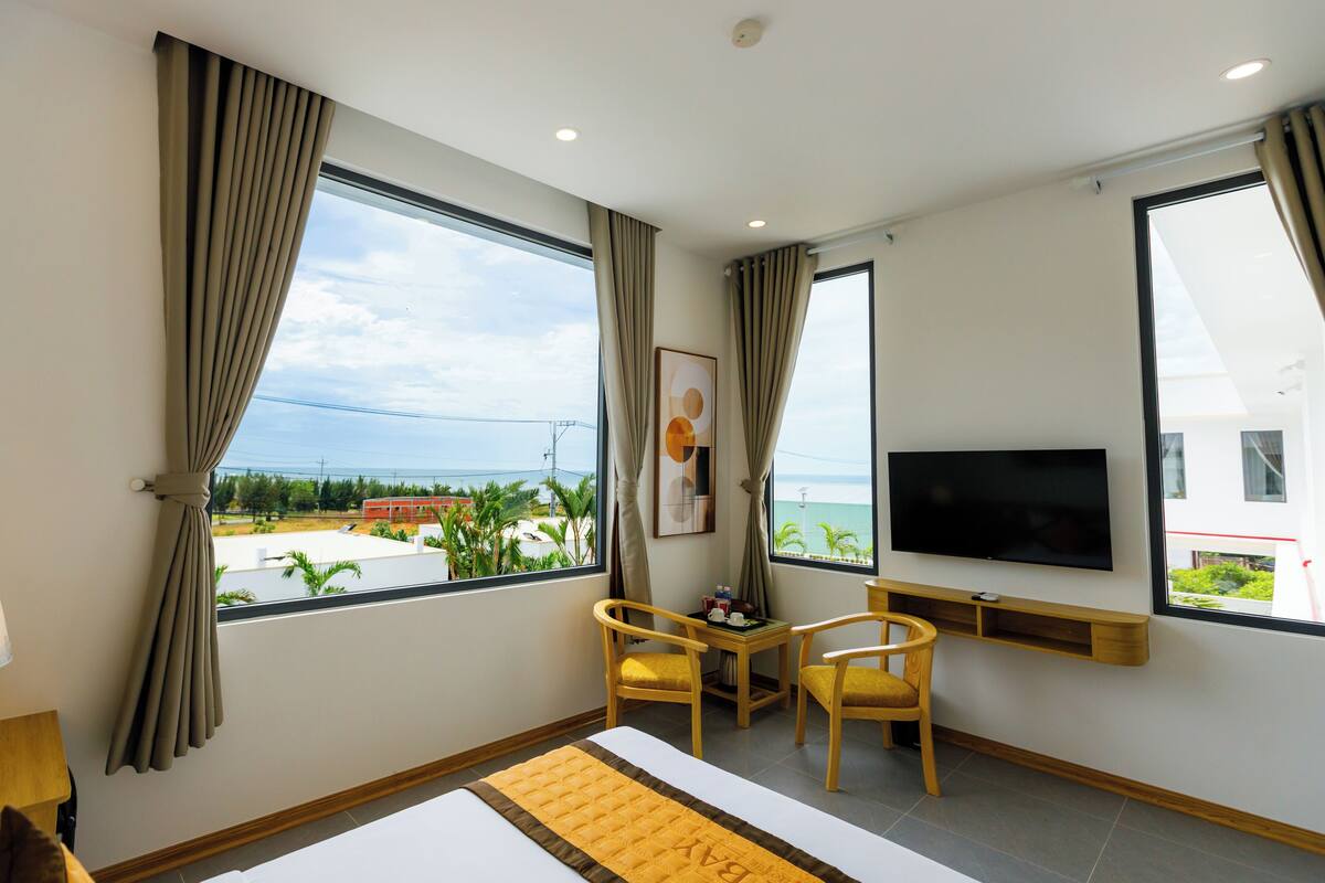 A hotel room with a bed, two chairs, a TV, and a window with a view of the sea.