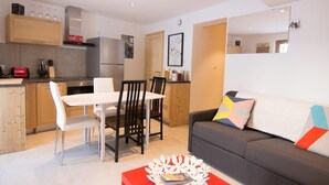 A modern kitchen with white cabinets, a built-in oven, and a refrigerator. There is a dining area with a white table and chairs, and a gray sofa with a colorful pillow.