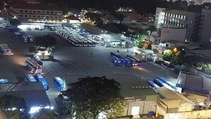 Vista nocturna de un estacionamiento con varios autobuses y automóviles, rodeado de edificios y árboles.