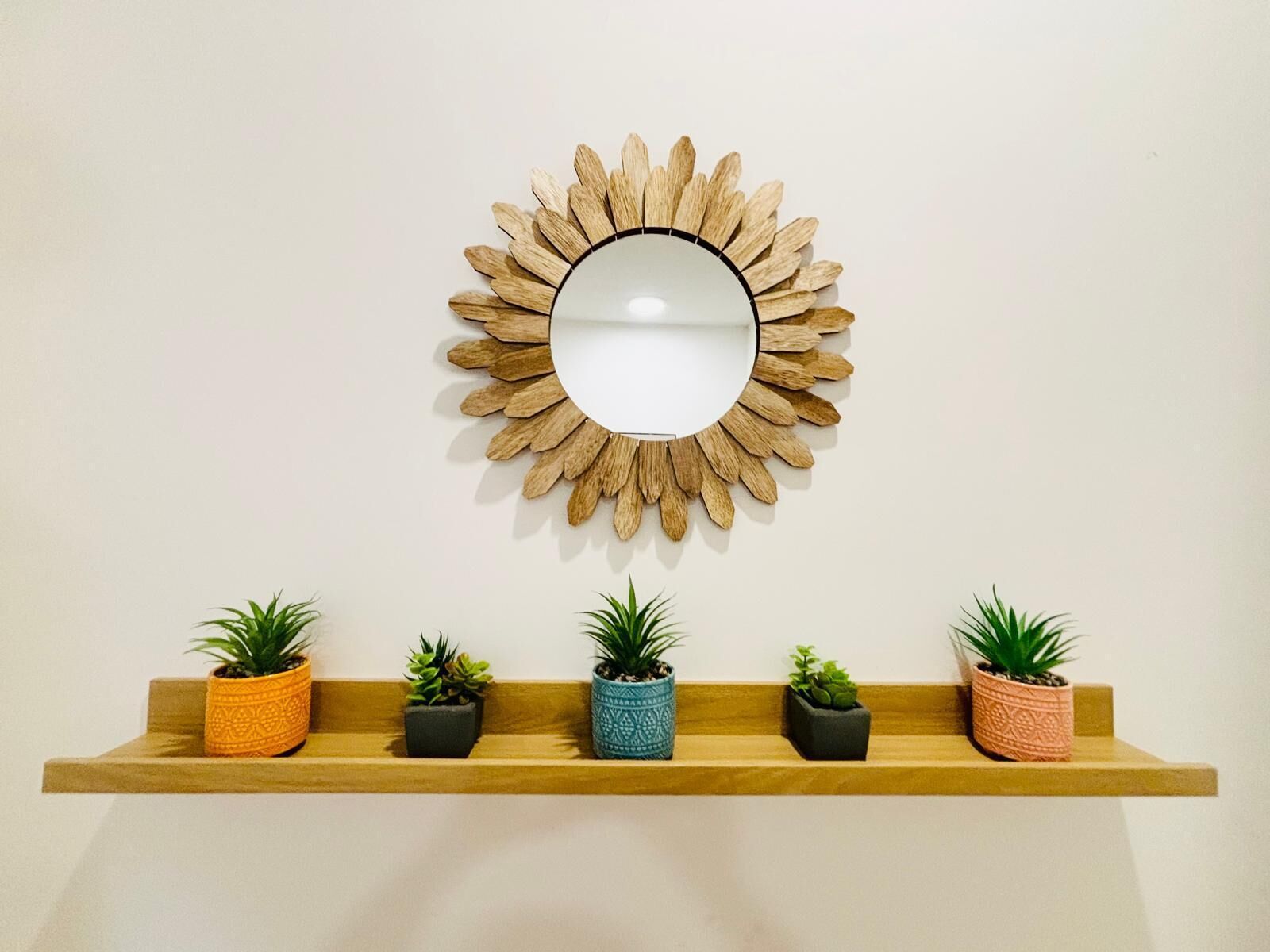A sunburst mirror on a wall with a wooden shelf below holding four potted plants.