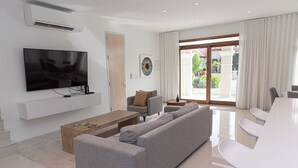 A modern living room with a flat-screen TV, grey sofa, coffee table, and a dining area with white chairs. - Aruba Opal Rental I (Noord)