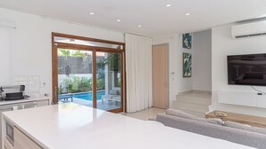 A modern living room with a large TV, a kitchen area, and a sliding glass door leading to a pool. - Aruba Opal Rental I (Noord)
