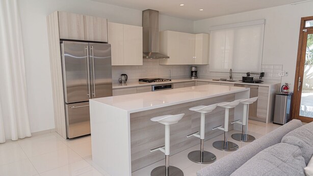 A modern kitchen with a central island, stainless steel appliances, and a grey sofa in the living area. - Aruba Opal Rental I (Noord)