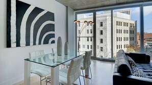 Una sala de estar moderna con una mesa de comedor de vidrio, sillas blancas, un sofá de cuero negro y un cuadro abstracto grande en la pared.