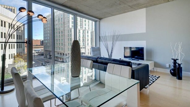 Una sala de estar moderna con una mesa de comedor de cristal, un sofá negro, un televisor y vistas a la ciudad a través de amplias ventanas.