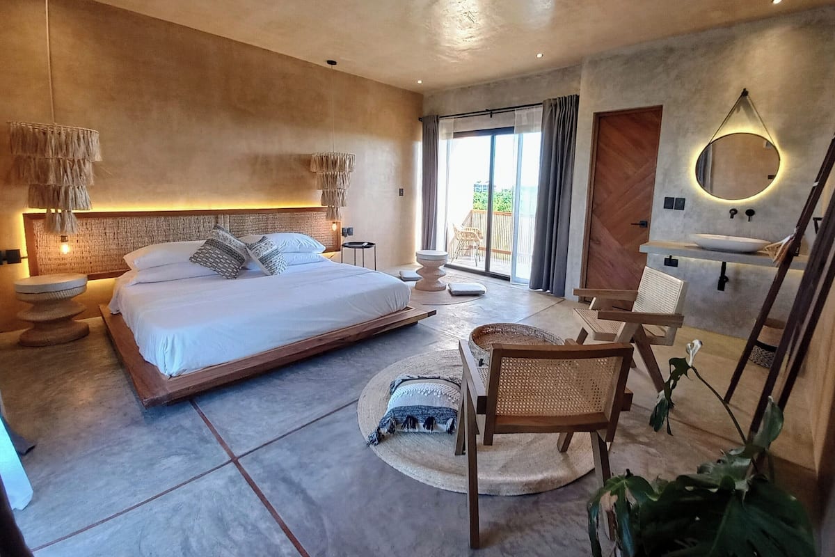 A modern bedroom with a large bed, a round mirror, a bedside table, and a view of the outdoors.