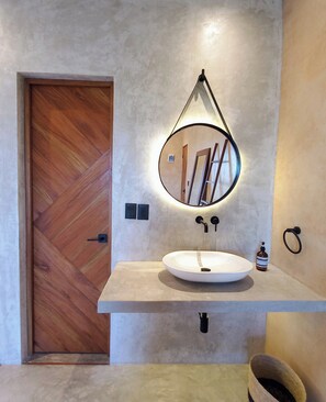A bathroom with a wooden door, a round mirror with a light, a white sink, and a towel ring. - Piquete Holbox (Isla Holbox)