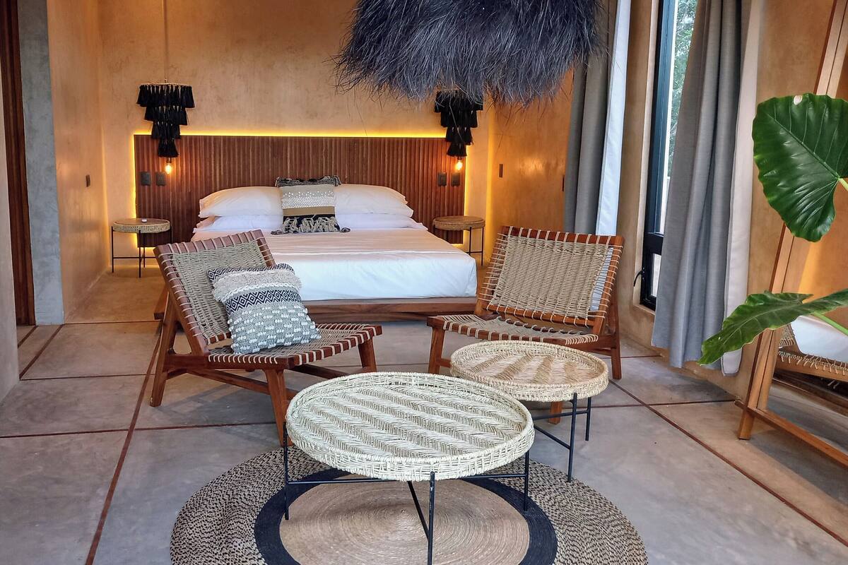 A modern bedroom with a large bed, two wicker chairs, a small round table, and a decorative feathered light fixture.