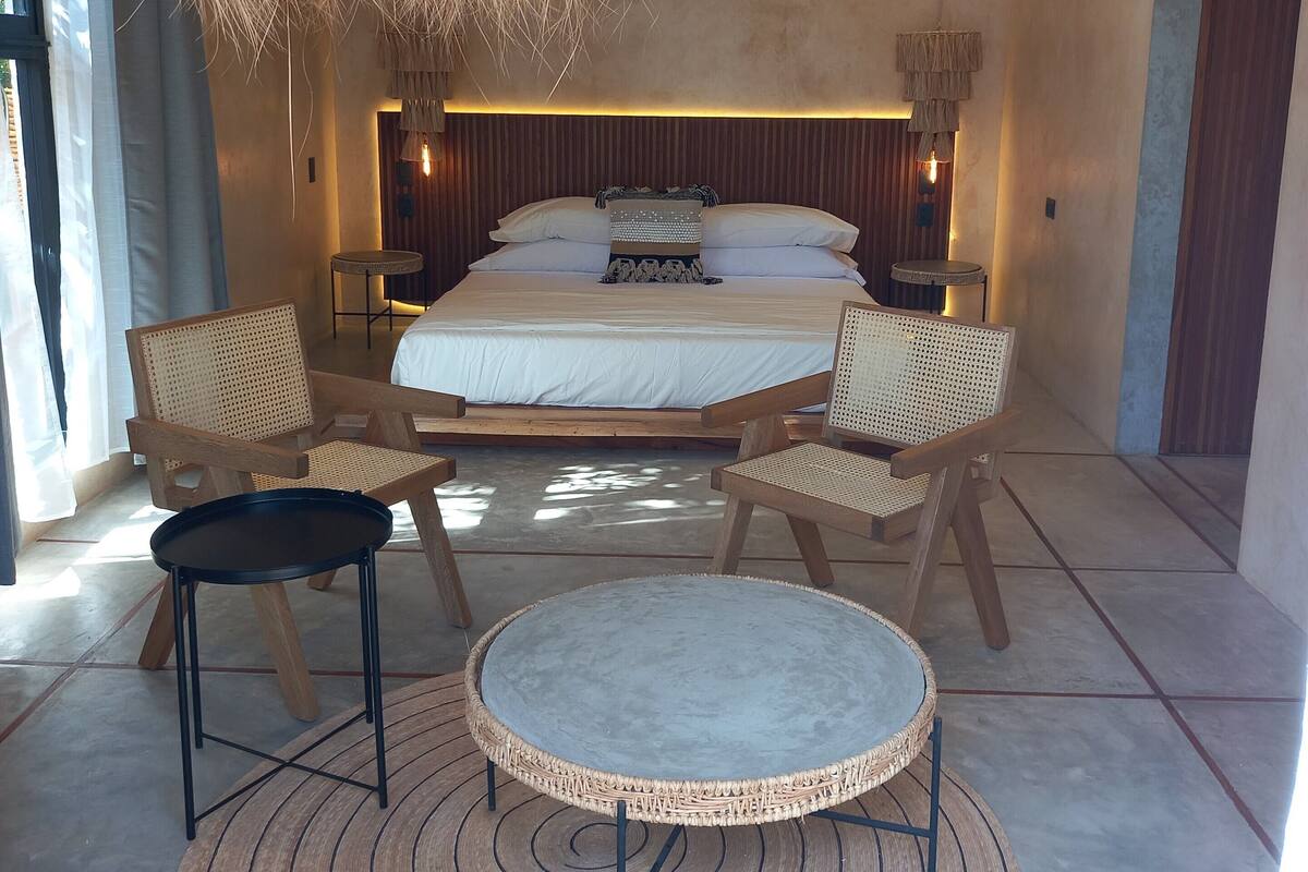 A modern bedroom with a large bed, two wooden chairs, a small round table, and a hanging light fixture.