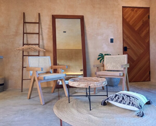 Quarto com duas cadeiras de madeira com assentos trançados, uma mesa redonda pequena, um tapete tecido e uma porta de madeira com padrão herringbone.