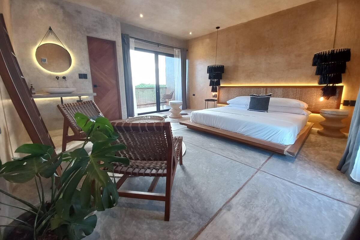 A modern bedroom with a large bed, a sitting area with a chair, a round mirror, and a view of the outdoors.