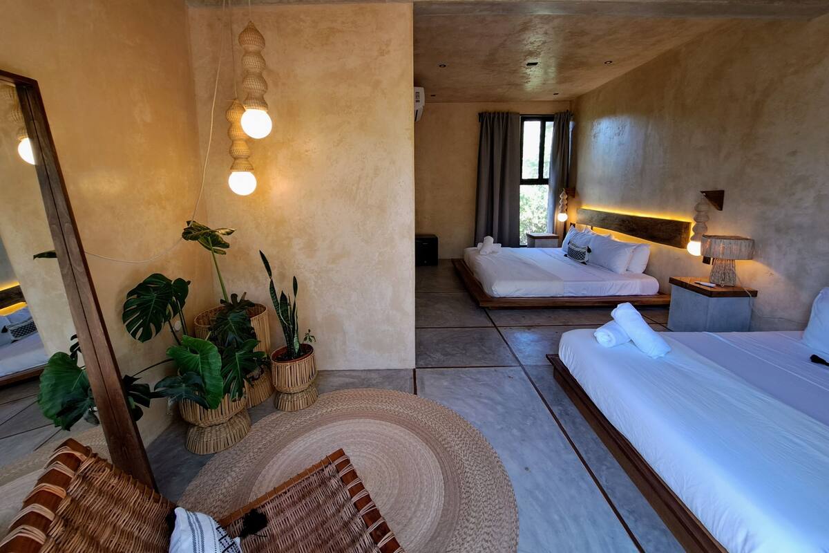 A modern hotel room with a large bed, a smaller bed, a sitting area with a chair, and a dining table. There are several potted plants and a large mirror.