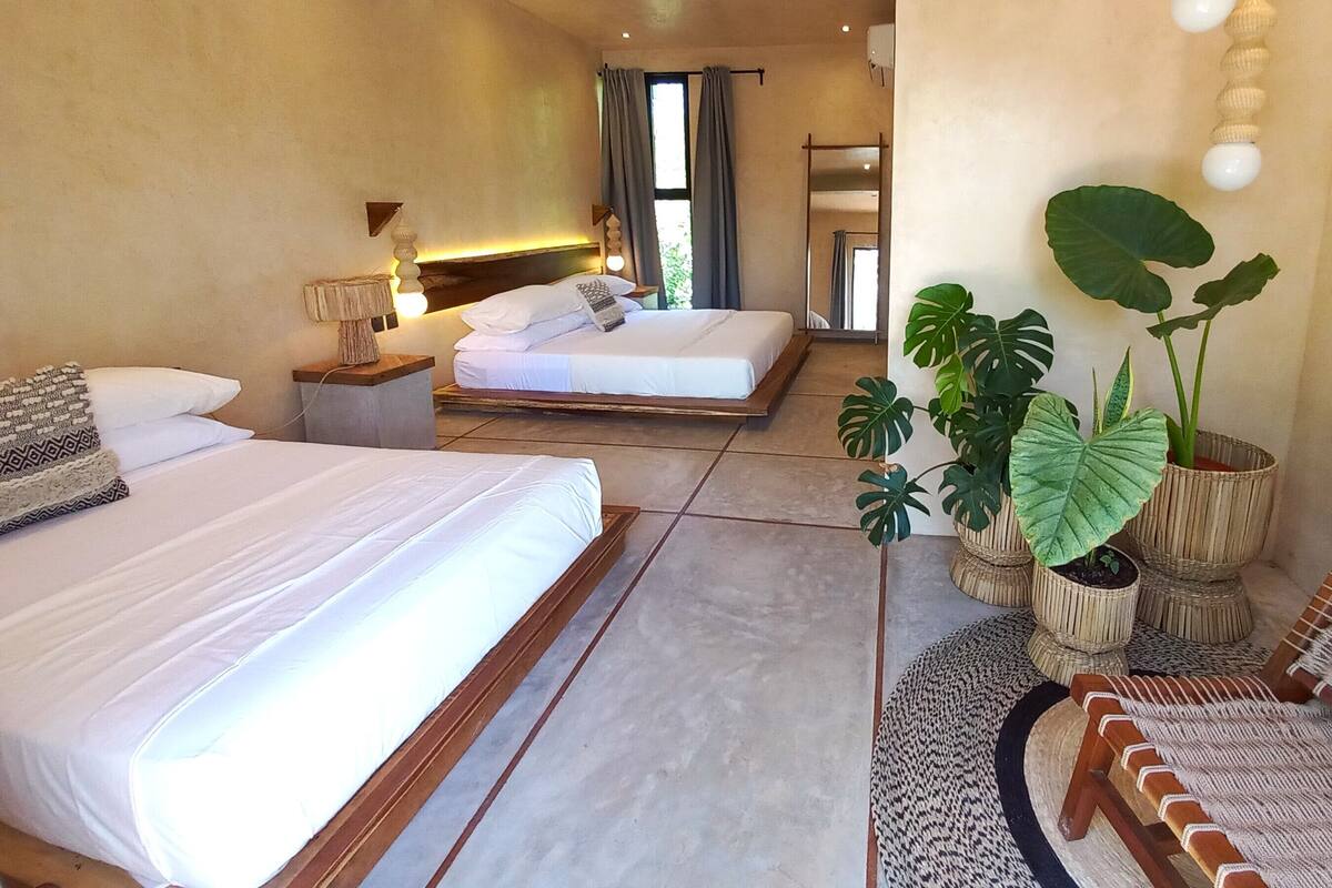 A hotel room with two beds, a large window with curtains, a bedside lamp, and a potted plant.