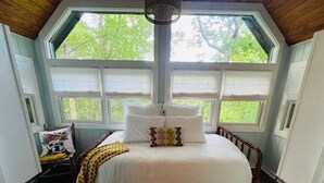 Un dormitorio con un ventanal grande, una cama con ropa de cama blanca, una silla de madera y una mesita de noche con una lámpara.