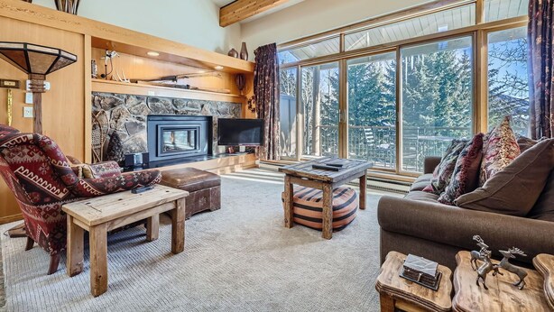 A cozy living room with a stone fireplace, large windows offering a view of trees, and rustic wooden furniture. - Stunning Third Story Condo with Stunning Mountain Views (Keystone)