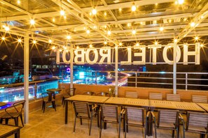 Un bar sul tetto con tavoli e sedie, vista piscina e lettere illuminate che compongono la scritta 'HOTEL'.