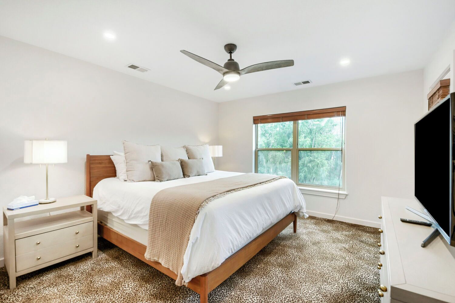 A bedroom with a bed, nightstands, a desk, a television, a ceiling fan, and a window with a view of trees.