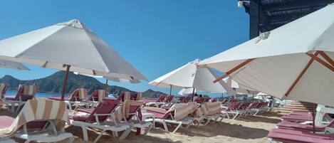 A beach with rows of sun loungers and umbrellas, a clear sky, and mountains in the background.