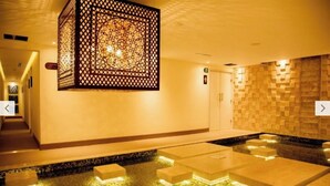 Un hall d’hôtel moderne avec un mur aux motifs géométriques, un comptoir d’accueil et un petit espace piscine doté de marches éclairées.