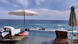 Une piscine à débordement offrant une vue sur l’océan, une chaise longue, une table et un parasol.