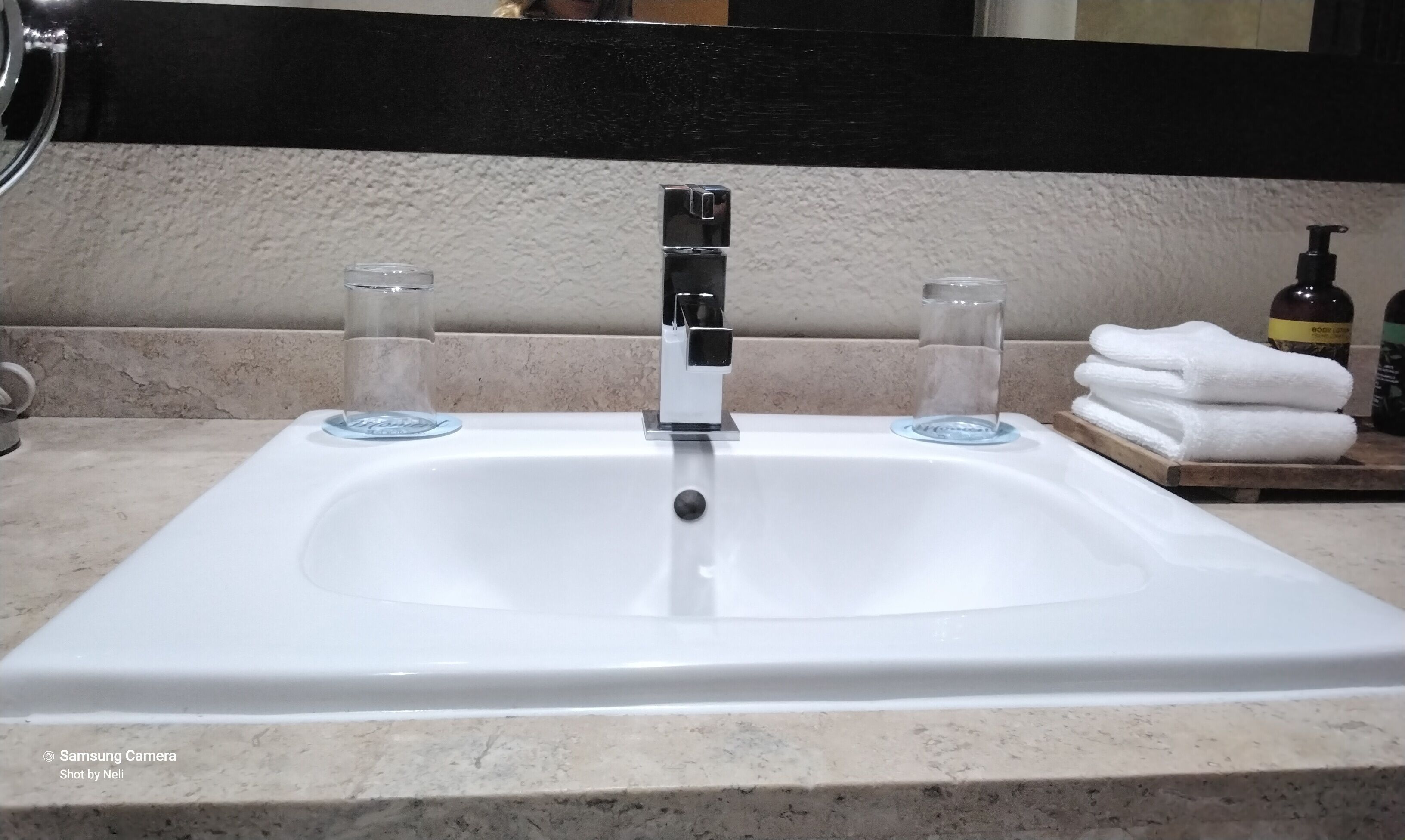 A bathroom sink with a chrome faucet, two clear glasses, and a wooden tray with folded towels.