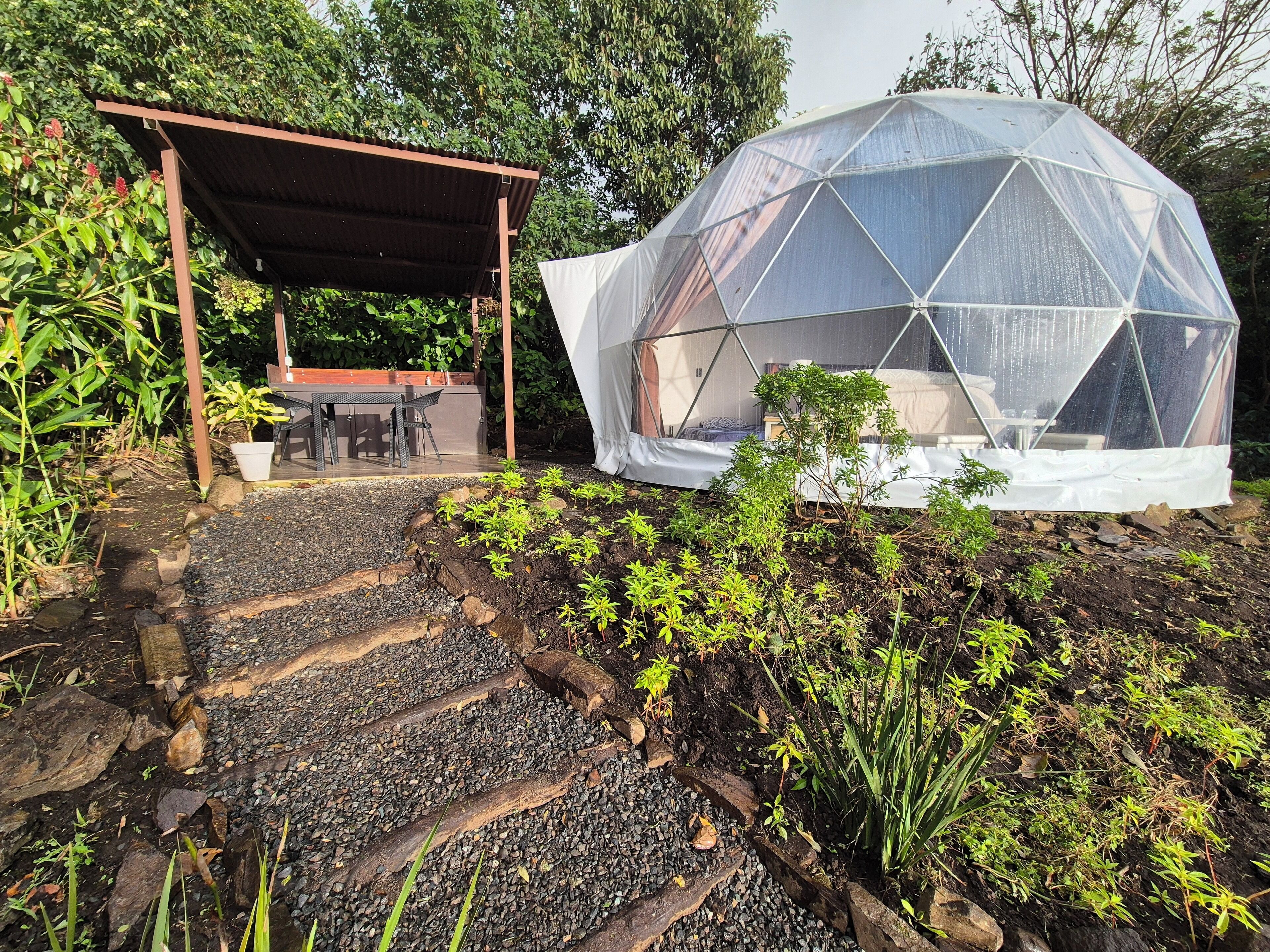 Une structure en forme de dôme géodésique dotée d’un espace extérieur aménagé avec des sièges et d’un sentier traversant un jardin.