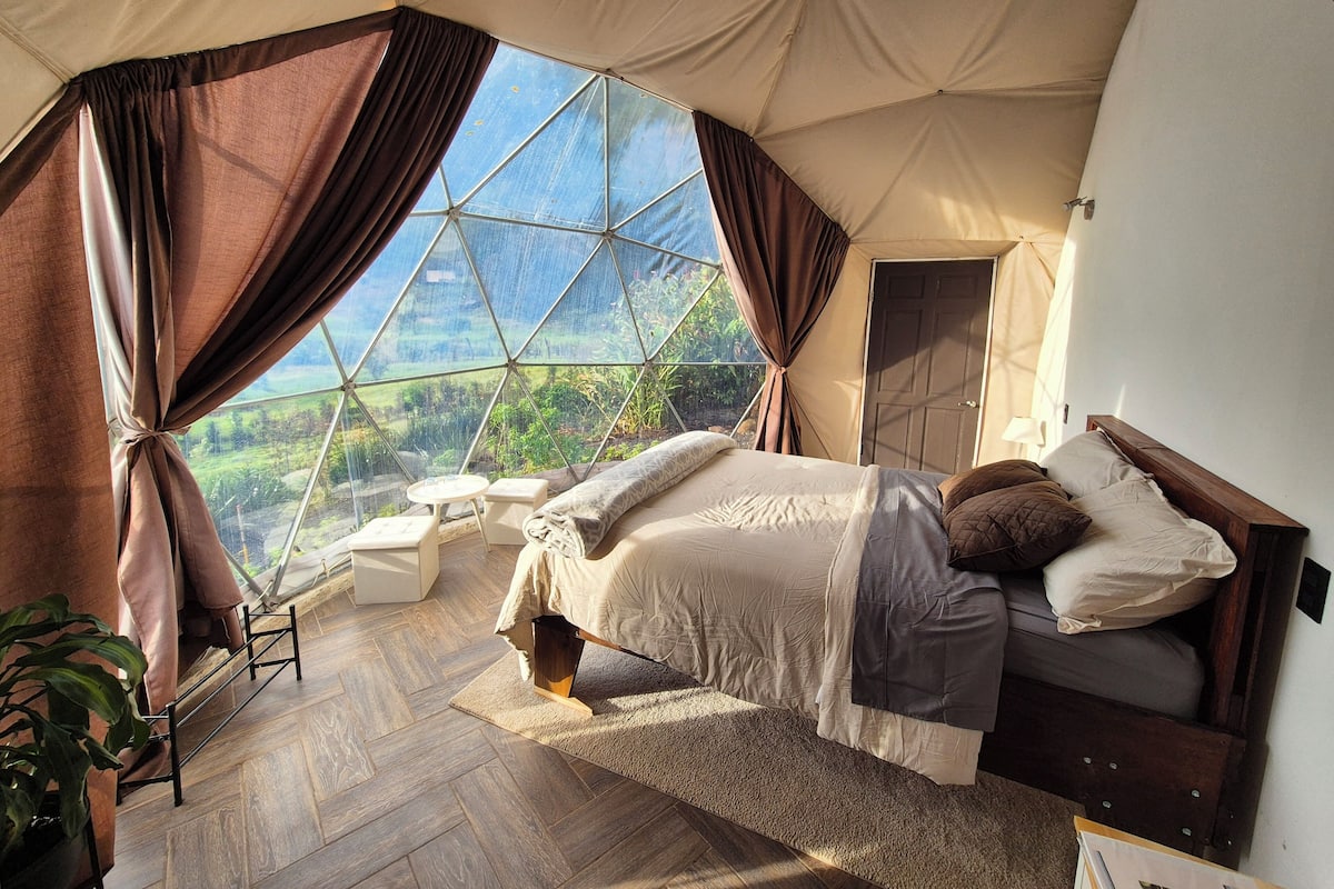 A bedroom with a large window, a bed, a bedside table, and a chair.