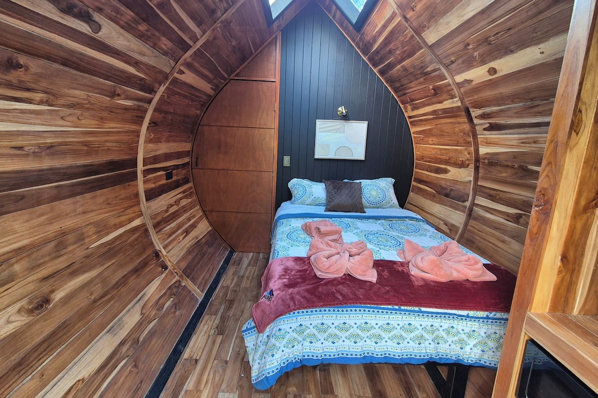 A cozy, circular wooden room with a bed, a wall-mounted light, and a framed picture.
