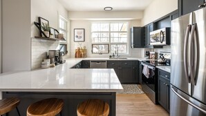 A modern kitchen with a white countertop, dark cabinets, stainless steel appliances, and wooden stools. - Tremblant Prestige - Les Eaux 233-3 (Mont-Tremblant)