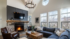 A living room with a stone fireplace, a leather armchair, a dark sofa, and a wooden coffee table. - Tremblant Prestige - Les Eaux 233-3 (Mont-Tremblant)