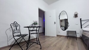 A room with a bed, a small table with a chair, a potted plant, a mirror, and a door leading to another room. - Maison Vaticana (Rome)