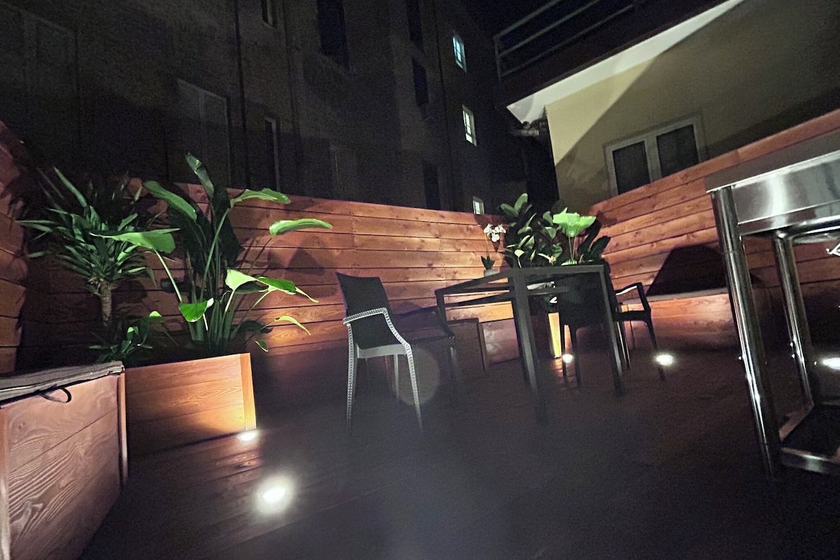 An outdoor patio area with wooden flooring, potted plants, a table with chairs, and a metal table.