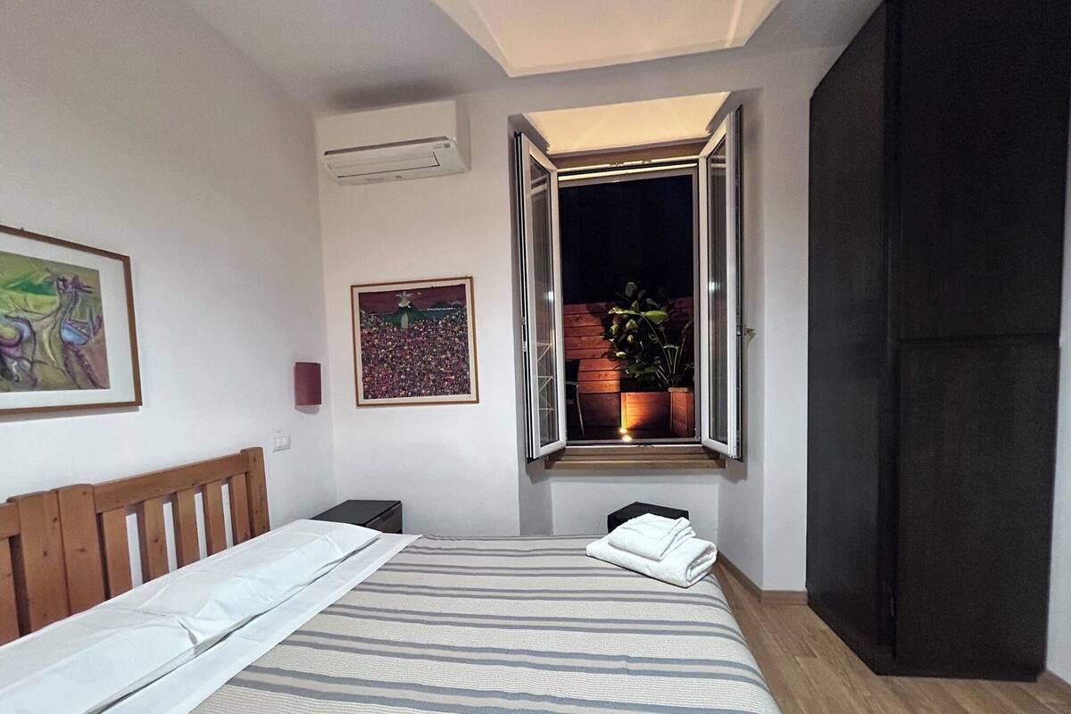 A bedroom with a bed, a window with a view of a garden, a painting on the wall, and an air conditioning unit.