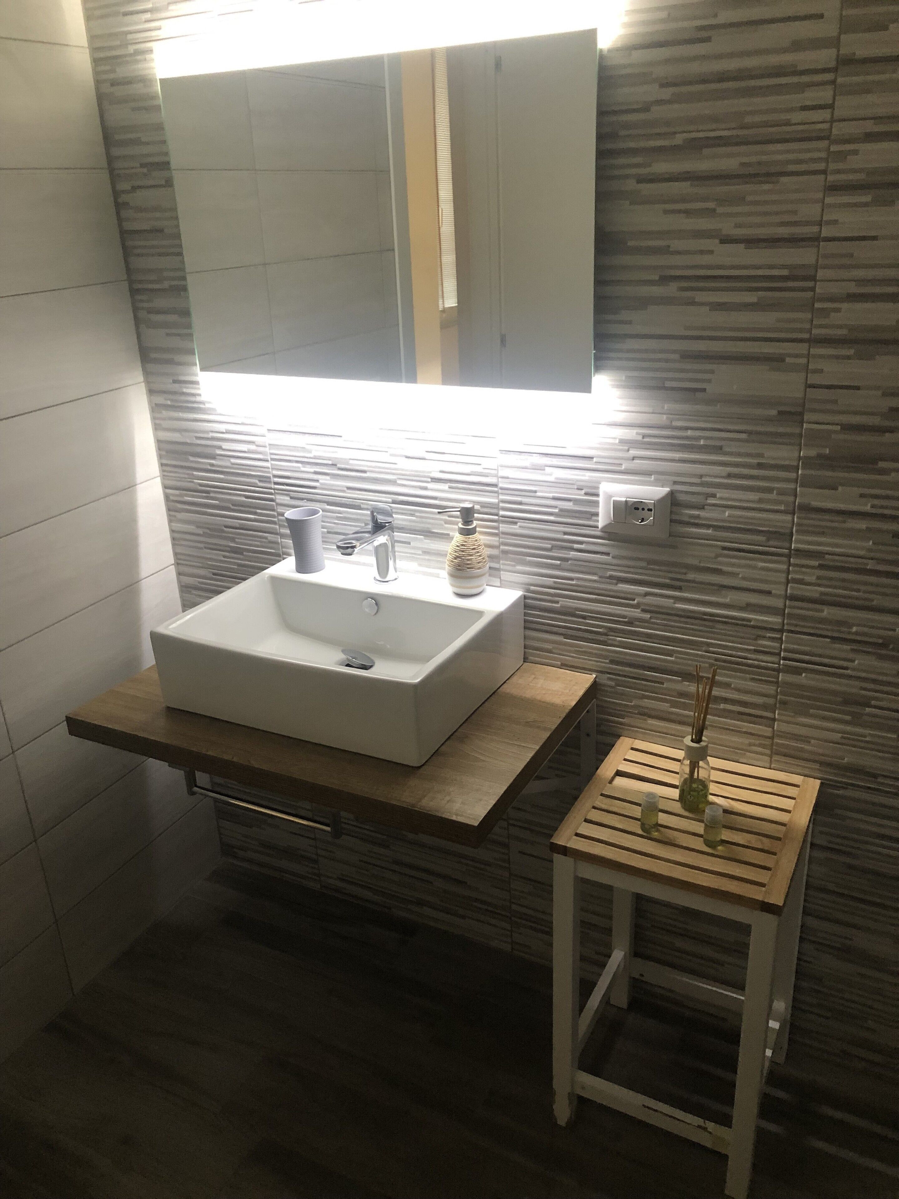 A modern bathroom with a rectangular sink, a mirror above it, and a small wooden side table with a bottle and two glasses.