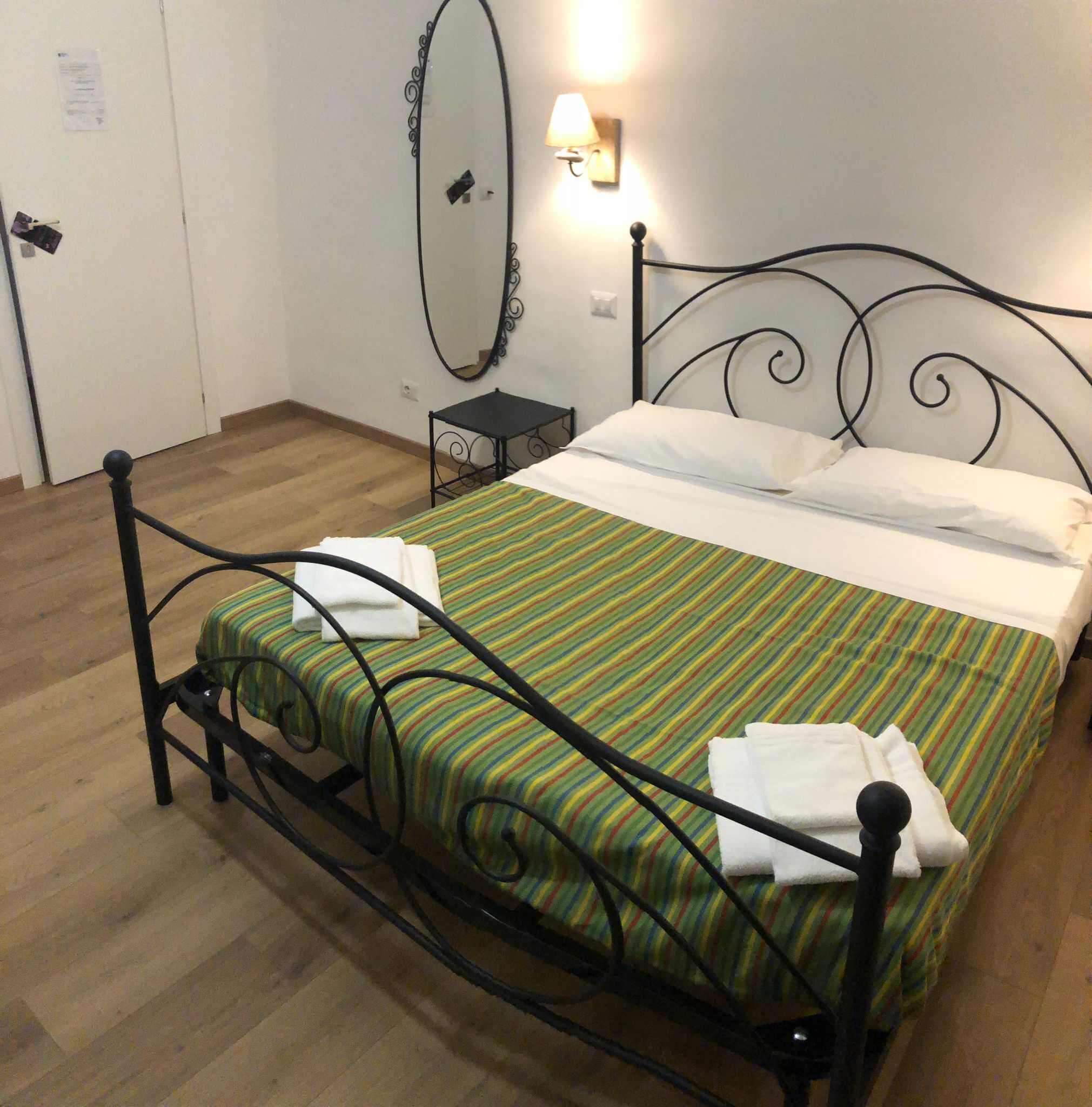 A single bed with a metal frame, a striped bedspread, and two pillows. There is a small bedside table with a lamp, a wall-mounted mirror, and a door with a paper on it.