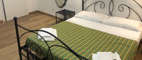 A single bed with a metal frame, a striped bedspread, and two pillows. There is a small bedside table with a lamp, a wall-mounted mirror, and a door with a paper on it.