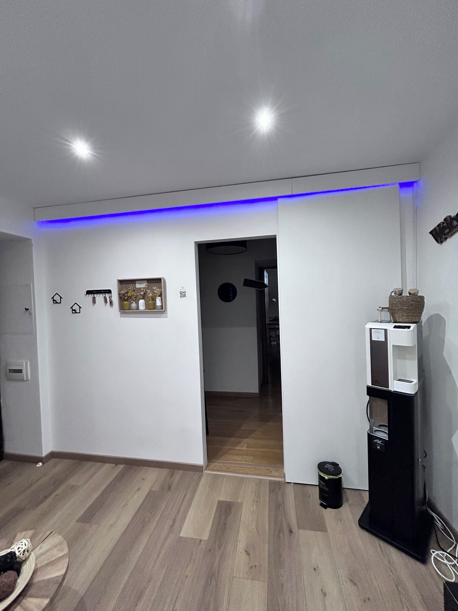 A modern room with a wooden floor, a water dispenser, and a blue LED light strip along the ceiling.