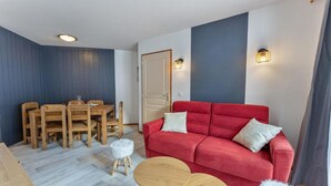 Ein Zimmer mit einem roten Sofa, einem Holzespeisetisch und Stühlen. Es gibt außerdem zwei kleine runde Tische und einen weißen, flauschigen Hocker.