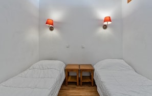 A small, single-room accommodation with two single beds, a wooden nightstand, and two wall-mounted lamps.