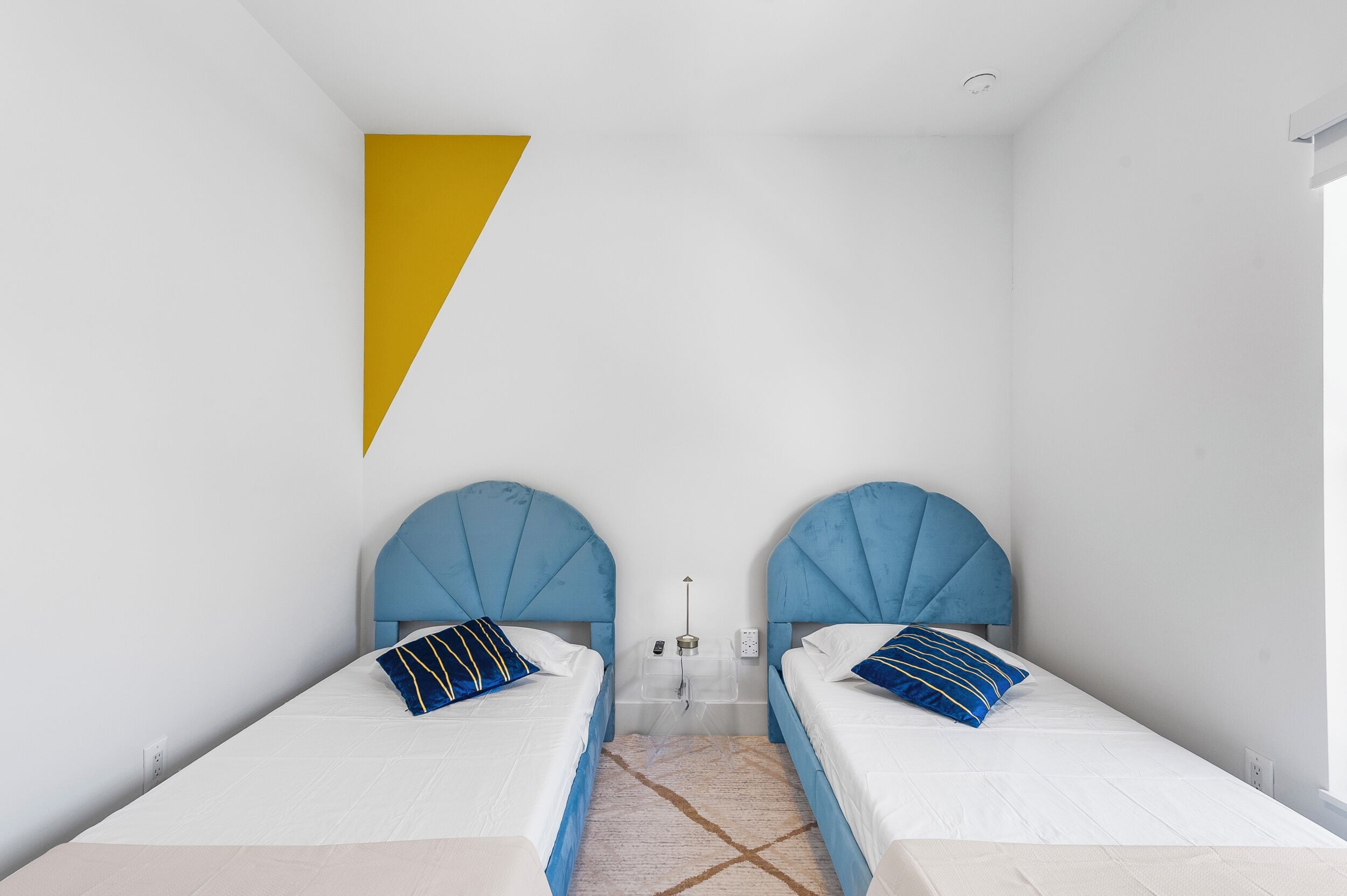 Een kamer met twee bedden, elk met een blauwe hoofdbord en een witte matras. Er is een gele, driehoekige decoratie aan de muur, een tafeltje met een lamp en een raam dat natuurlijk licht biedt.