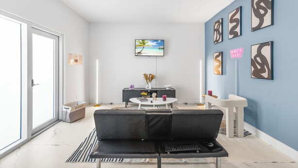 A modern living room with a black sofa, a round coffee table, and a TV mounted on the wall. There is a sliding glass door on the left and a blue accent wall with abstract art. - Modern 3BR Near Beaches–Fast WiFi & Spacious Design (Fort Lauderdale)