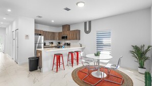 A modern kitchen with a central island, red bar stools, a round table with chairs, and a potted plant. - Modern 3BR Near Beaches–Fast WiFi & Spacious Design (Fort Lauderdale)