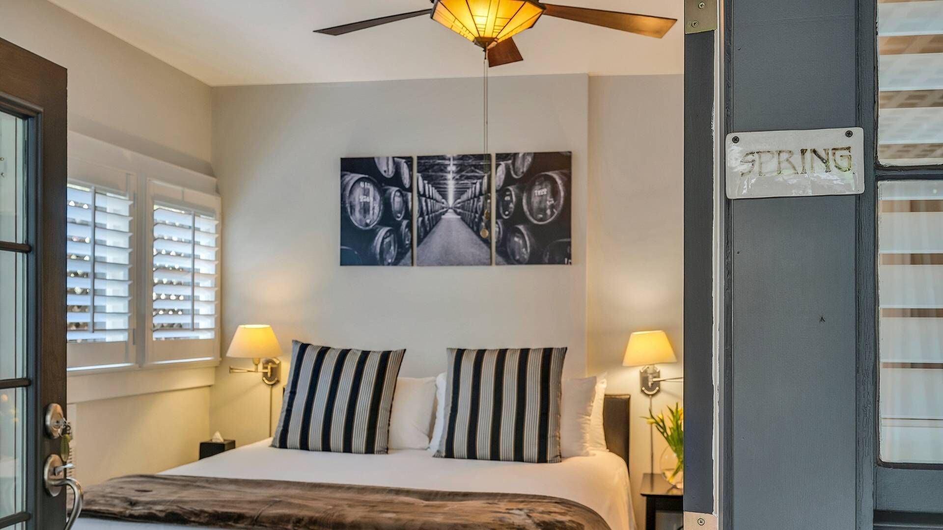 Une chambre d’hôtel avec un lit, deux lampes de chevet, un ventilateur de plafond, une œuvre d’art accrochée au mur et une pancarte sur la porte.