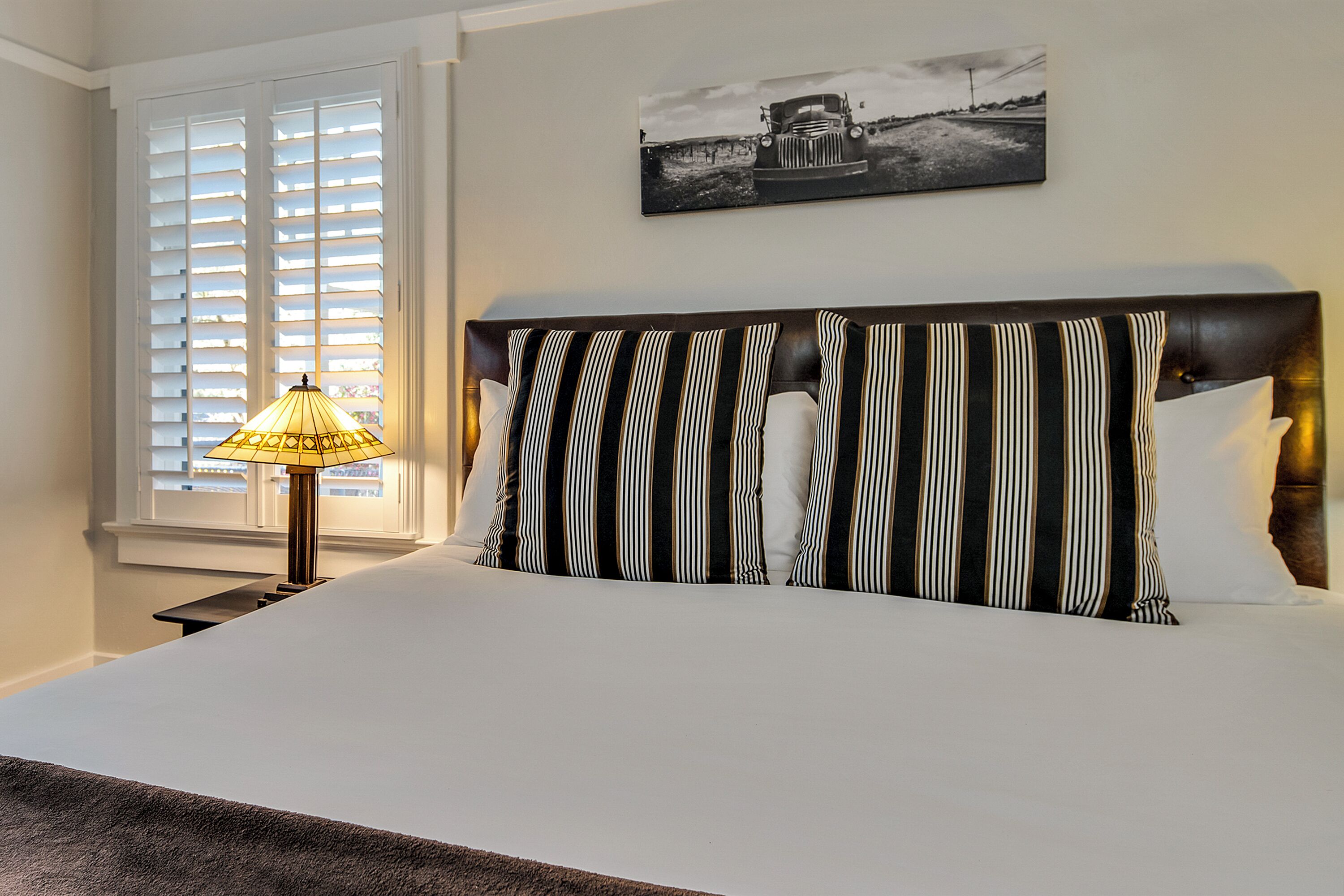 A neatly made bed with striped bedding, a lamp on a bedside table, and a black and white photograph on the wall.