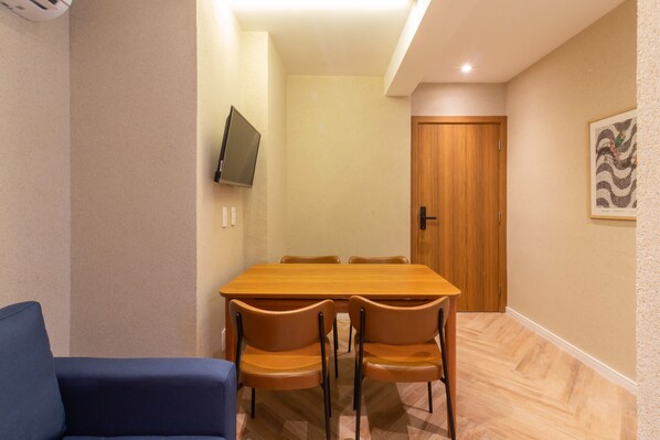 Una habitación con una mesa de comedor de madera, cuatro sillas, un sofá azul, un televisor montado en la pared y una puerta de madera.
