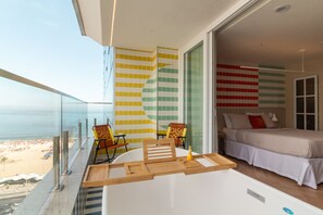 Un balcón con vistas a la playa, una cama con cubrecama blanco y una pared con un colorido diseño de azulejos.