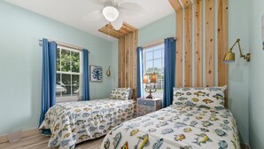 A bedroom with two beds, wooden ceiling beams, a ceiling fan, a nightstand with a lamp, and a window with curtains.