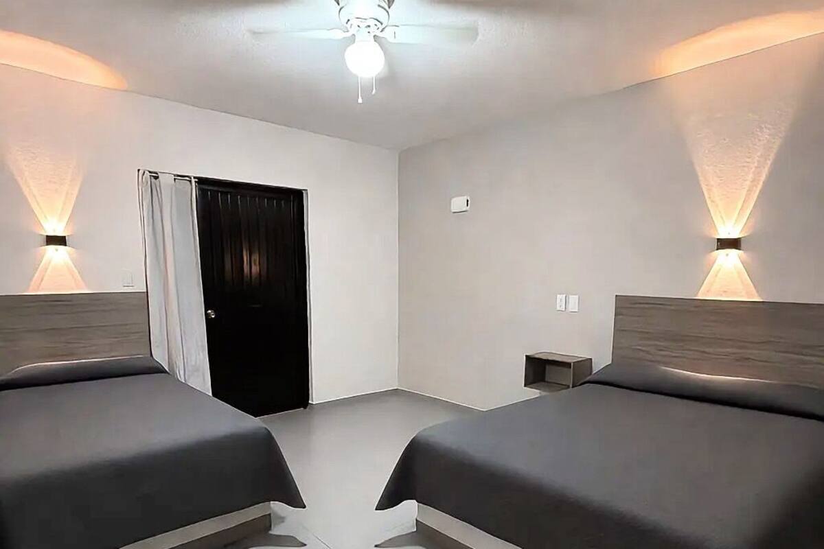 Habitación de hotel con dos camas, ventilador de techo, una puerta e iluminación en la pared.