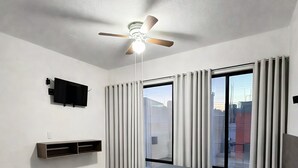 Un dormitorio con cama, ventilador de techo, televisor y ventanas amplias con cortinas.
