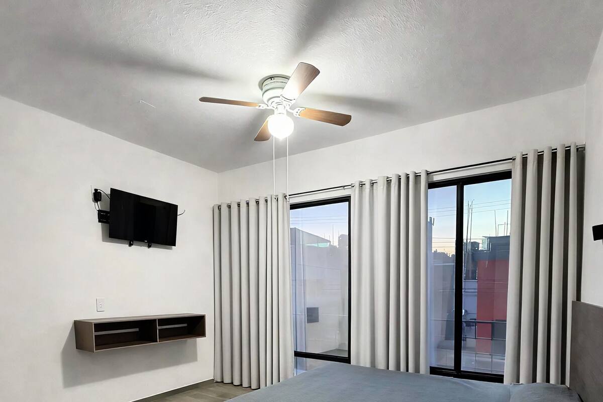 Un dormitorio con cama, ventilador de techo, televisor y ventanas amplias con cortinas.